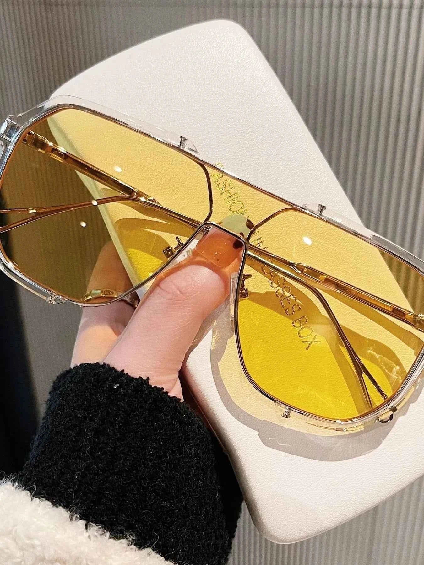 Trendy gold-tone aviator sunglasses with yellow lenses, held over a white case, perfect for fashion accessories.