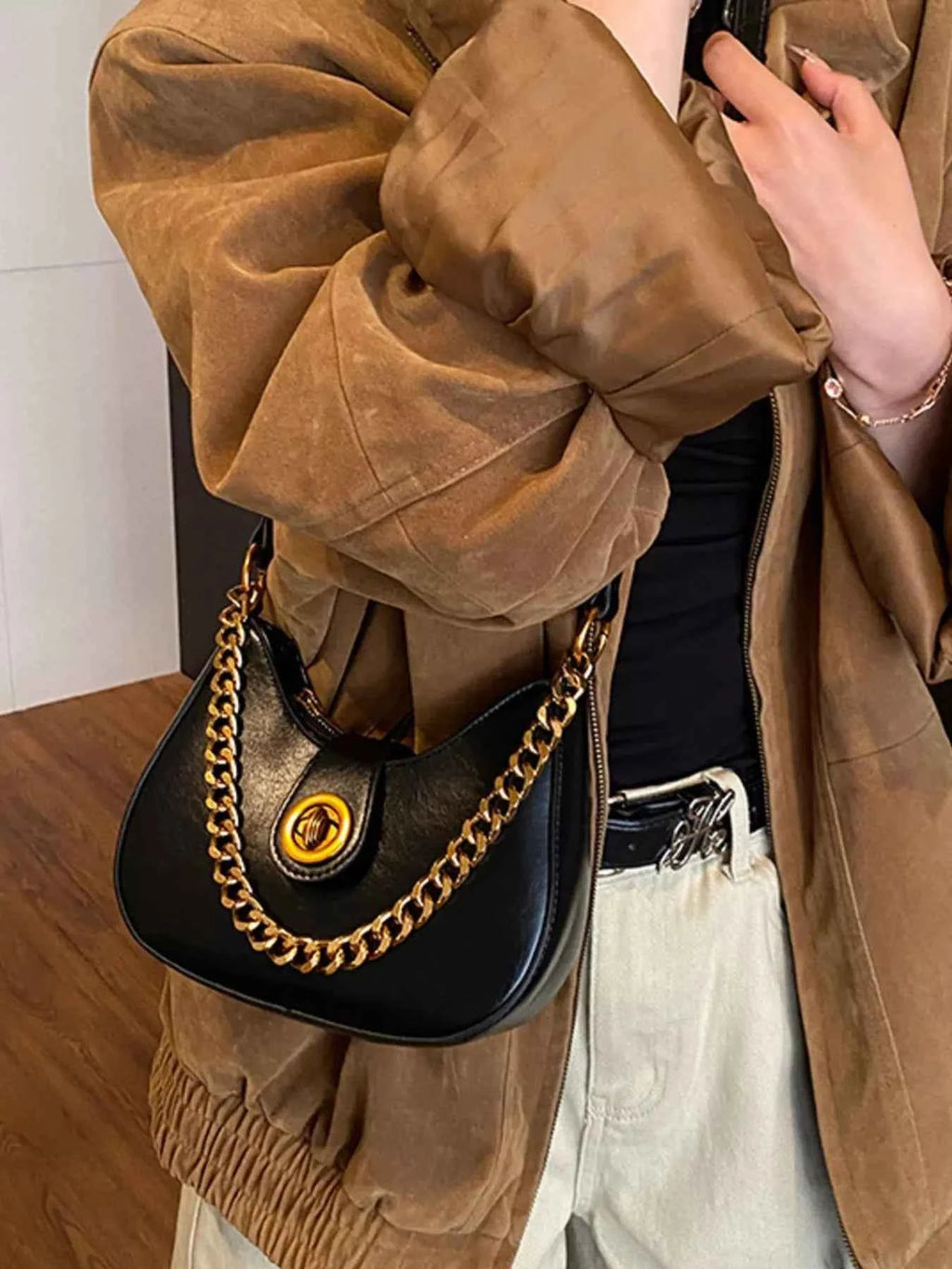 Stylish black crescent bag with gold chain strap, worn with brown jacket and beige pants.