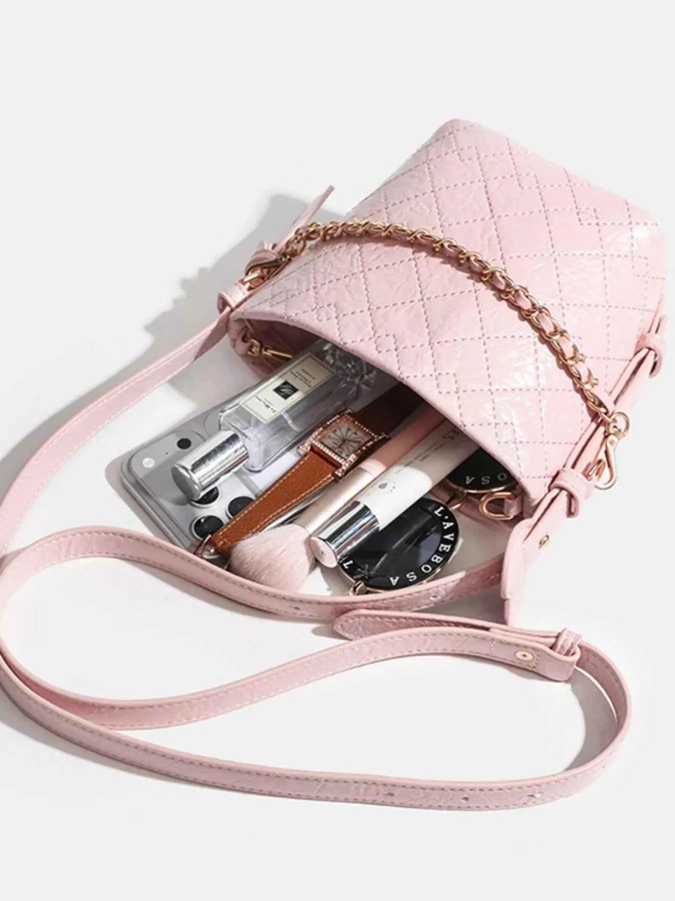 Pink quilted crossbody bag with gold chain strap, holding phone, perfume, makeup, and accessories