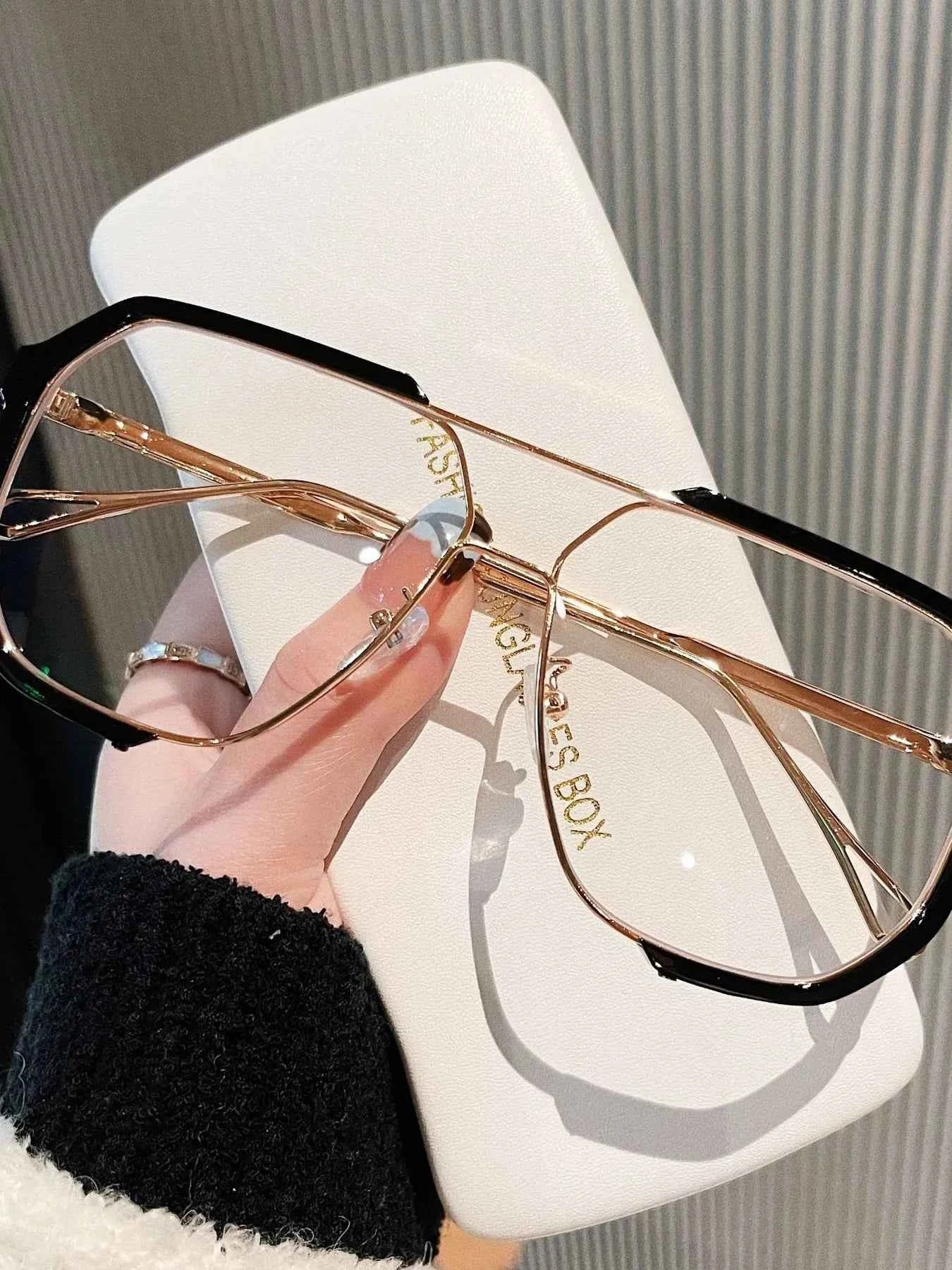 Trendy oversized glasses with black and gold frames, held over a white case, perfect for fashion accessories.