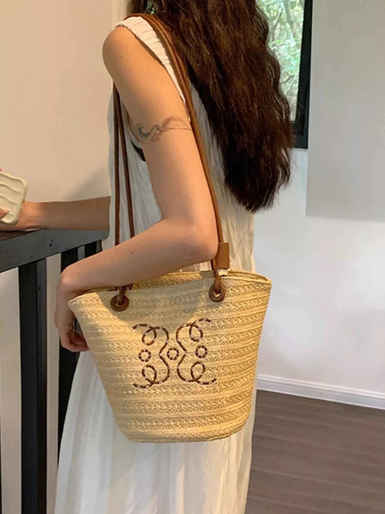 Straw tote bag with brown leather straps and monogram, carried by woman in white dress