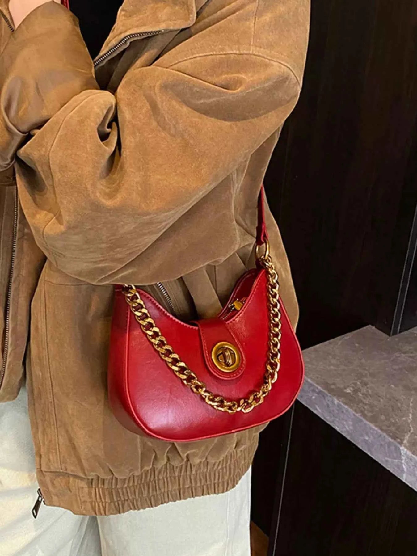 Red crescent-shaped handbag with gold chain strap, worn with brown suede jacket for trendy street style.