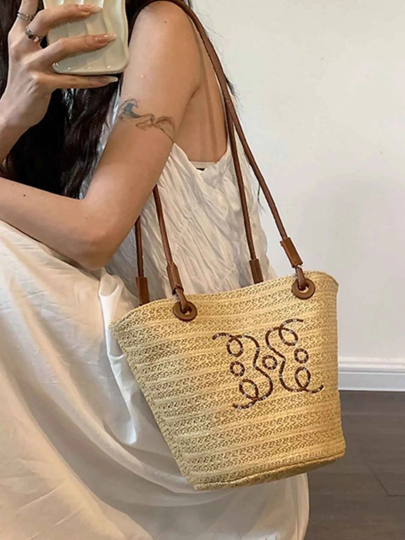 Woven straw tote bag with brown leather straps and decorative monogram, styled with white dress for summer fashion.