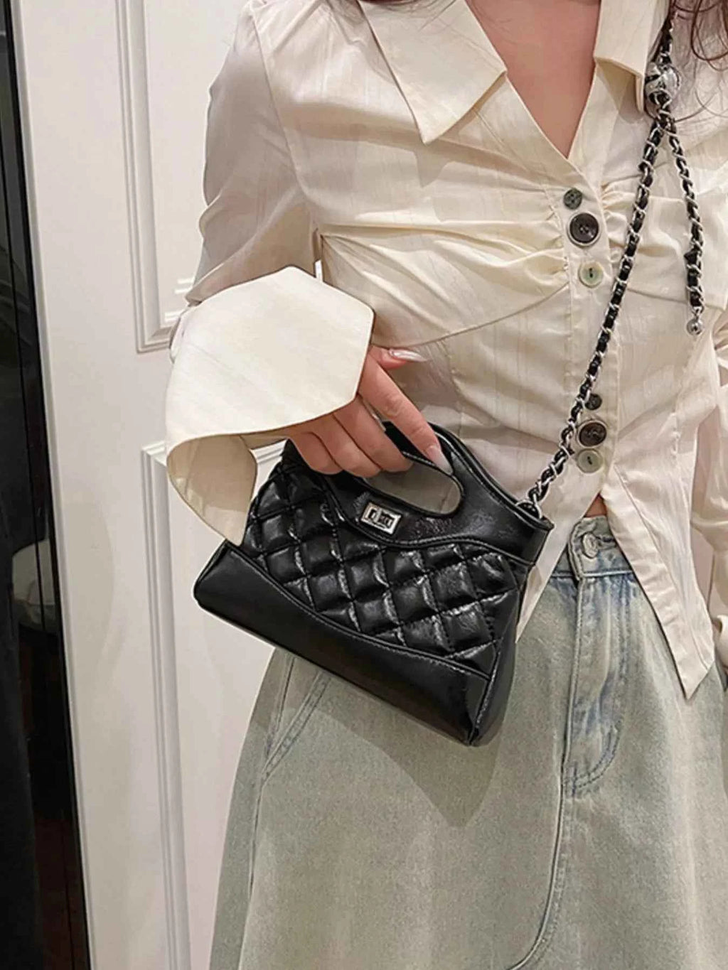 Black quilted handbag with chain strap, held by woman in white blouse and denim skirt