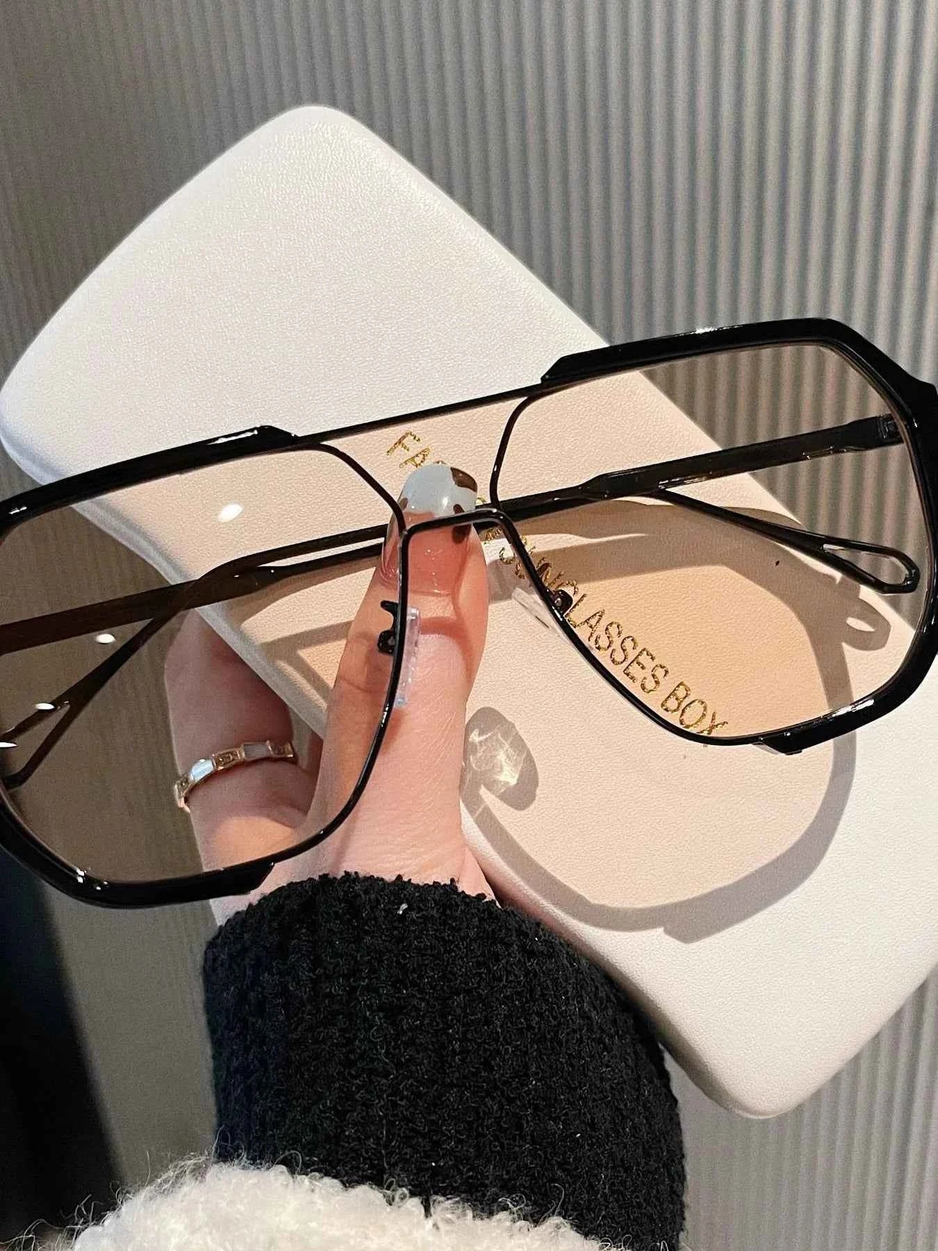 Oversized black frame sunglasses with clear lenses, held over white case, trendy fashion accessory